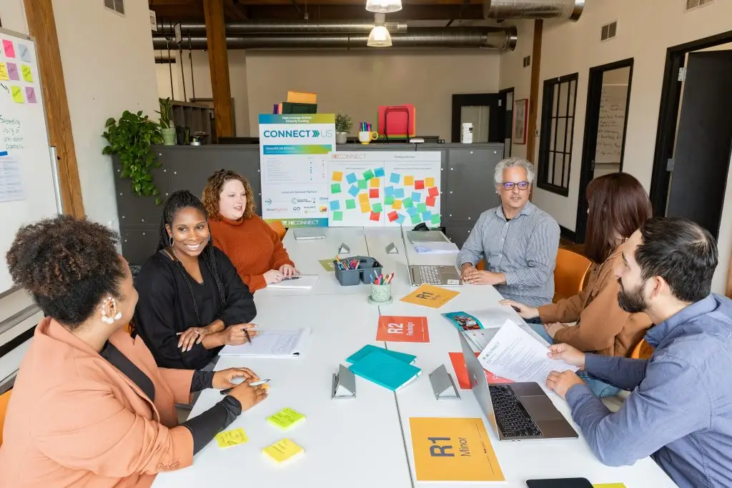 Diverse team at non-profit youth organization collaborating with sticky notes and laptops. "CONNECT" banner visible on planning board in modern workspace.