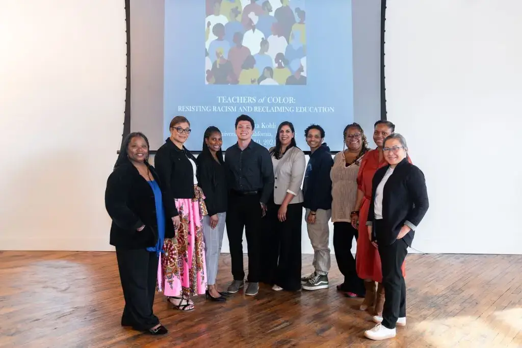 A diverse group of educators stands together at a "Teachers of Color" event focused on resisting racism and reclaiming education. The professionally dressed participants pose in front of a projection screen in a bright community space, embodying educational leadership and community partnership action for educational equity.