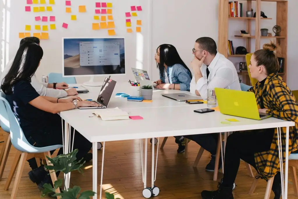 Diverse team collaborating at a conference table during a systemic change workshop, with colorful sticky notes on the wall documenting ideas and a digital presentation visible on screen.