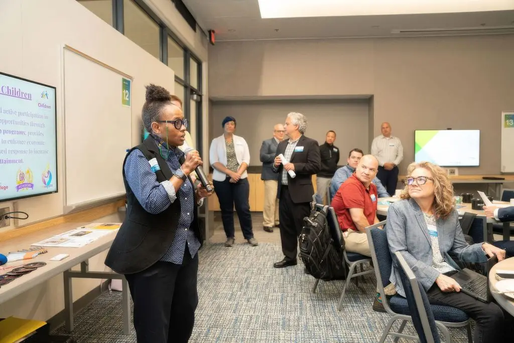 A woman speaks into a microphone during a meeting focused on systemic change, with participants seated and standing around the room.