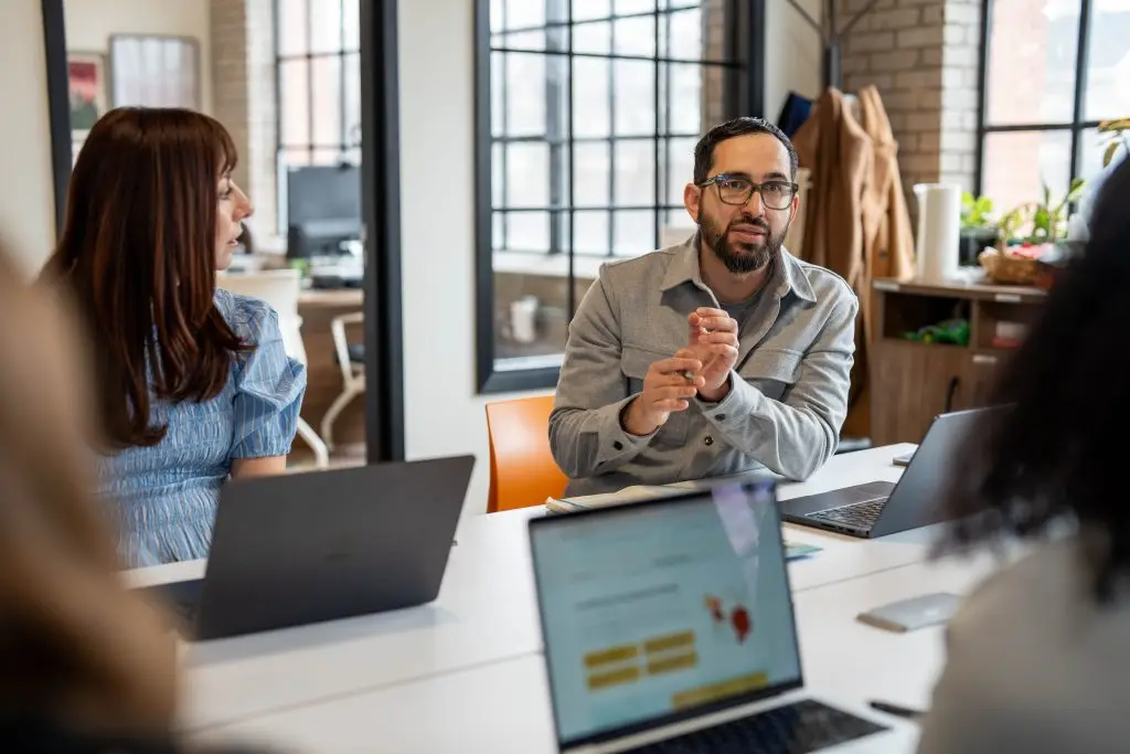 Salvador talks to the team with his hands clasped. Team members look at him with computers on the table near them.